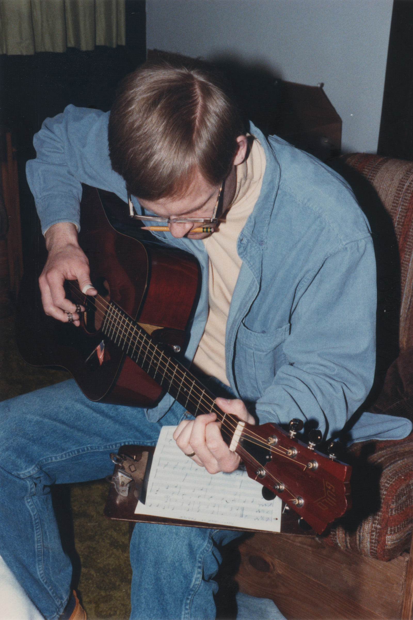 American songwriter Bill Schachter – pencil clenched in teeth, playing guitar with fingerpicks while reading handwritten sheet music.